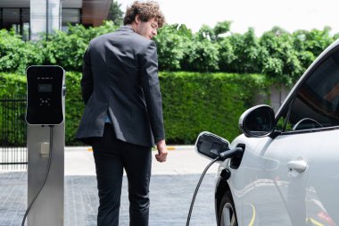 Modern şehir yerleşim bölgesindeki kamu şarj istasyonunda elektrikli araba şarj eden yenilikçi bir işadamı. Çevre dostu şarj edilebilir araba alternatif temiz enerjiyle çalışıyor..