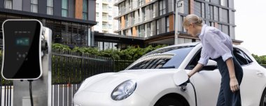 Progressive businesswoman insert charger plug from charging station to her electric vehicle with apartment condo building in background. Eco friendly rechargeable car powered by sustainable energy.