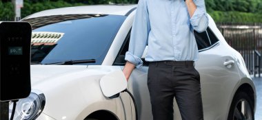 Closeup progressive suit-clad businessman with his electric vehicle recharge his car on public charging station in modern city with power cable plug and renewable energy-powered electric vehicle.