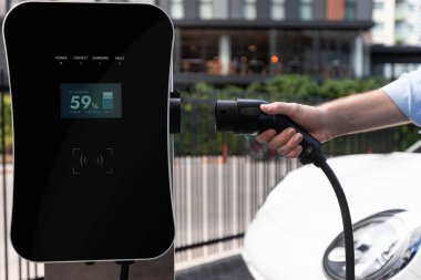 Closeup progressive man holding EV charger plug from public charging station for electric vehicle with background of residential building as concept eco-friendly sustainability energy car concept.