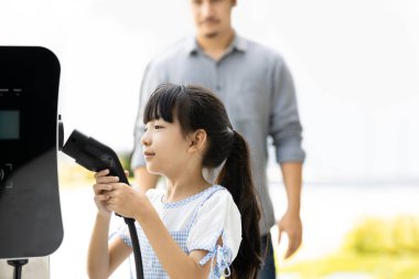 Focus progressive young girl holding EV charger device from home electric charging station for renewable energy powered car with blur asian man background. Sustainable green energy technology concept.