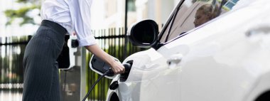 Closeup progressive suit-clad businesswoman with her electric vehicle recharge her car on public charging station in modern city with power cable plug and renewable energy-powered electric vehicle.