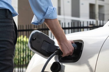 Closeup progressive man holding EV charger plug from public charging station for electric vehicle with background of residential building as concept eco-friendly sustainability energy car concept.
