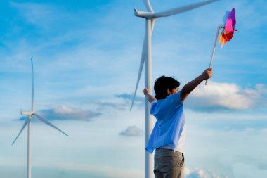 Progressive young asian boy playing with wind pinwheel toy in the wind turbine farm, green field over the hill. Green energy from renewable electric wind generator. Windmill in the countryside concept
