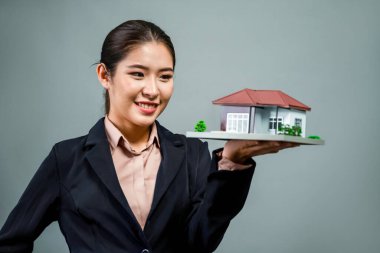 Resmi takım elbise giyen genç Asyalı kadın izole edilmiş arka planda ev modelini tutuyor ve sergiliyor. Konut ilanı, ev sigortası promosyonları ya da kişiselleştirilebilir fotokopi alanı için ideal. Coşkulu.
