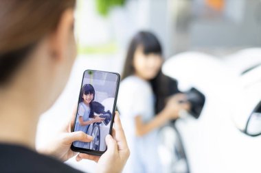 Akıllı telefon kullanan ilerici anne, EV 'yi şarj eden kızın odaklı fotoğrafını çekiyor. Asyalı bir kızın görüntüsü bulanık arkaplanlı telefon ekranında ev şarj istasyonundan EV vagonuna elektrikli şarj cihazı takıyor.