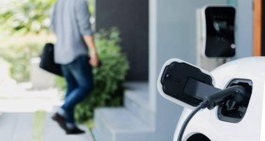 Focus electric car charging at home charging station with blurred progressive man walking in the background. Electric car using renewable clean for eco-friendly concept.