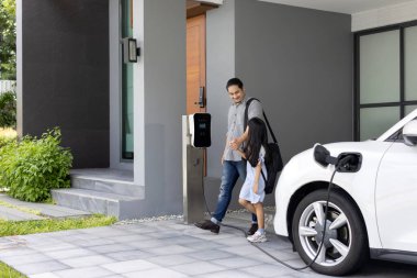 Progressive father and daughter plugs EV charger from home charging station to electric vehicle. Future eco-friendly car with EV cars powered by renewable source of clean energy.