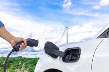 Progressive natural scenic with windmill generator where hand insert charging plug to electric vehicle from charging station with natural background. EV car powered by wind turbine electric generator.