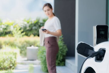 Focus image of electric vehicle recharging battery at home charging station with blurred woman walking in the background. Progressive concept of green energy technology applied in daily lifestyle.