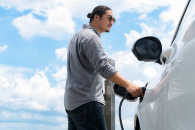 Progressive man with electric vehicle and cloudscape background. EV car driven by clean renewable plugged-in with charging point. Refreshing environment from green energy.
