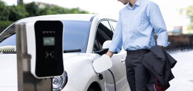 Closeup progressive man holding EV charger plug from public charging station for electric vehicle with background of residential building as concept eco-friendly sustainability energy car concept.