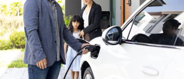 Progressive young parents and daughter with electric vehicle and home charging station. Green and clean energy from electric vehicles for healthy environment. Eco power from renewable source at home.