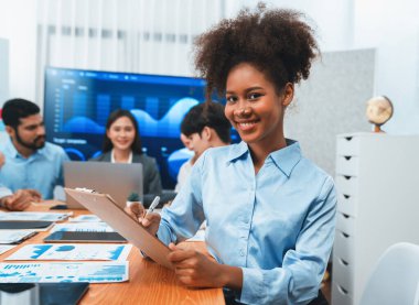 Bir grup ofis çalışanı ile birlikte arka planda ekranda gösterim paneliyle buluşan mutlu Afrikalı genç bir iş kadını portresi. Takım toplantısında kendine güveni tam bir ofis hanımı. Uyum