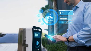 İş adamı EV akıllı telefon uygulamasından batarya durum hologramını kontrol ederken EV arabası da konut alanındaki şarj istasyonundan şarj oluyor. Fütürist temiz sürdürülebilir EV teknolojisi. İnceleyin