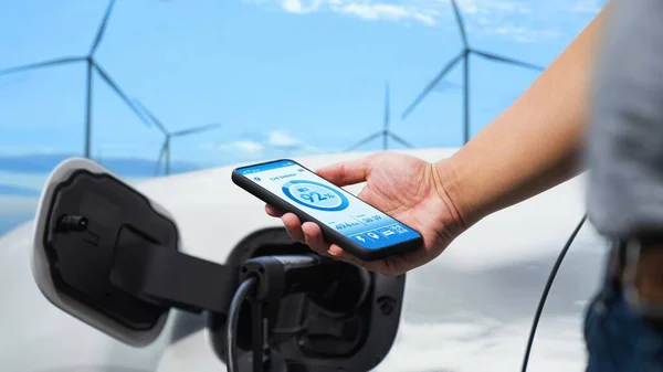 ビジネスマンは 風力発電機からの代替クリーンエネルギーを使用して充電ステーションから環境に優しいEv車を充電しながら スマートフォンのアプリケーションにバッテリーステータスディスプレイを確認します Peruse — ストック写真