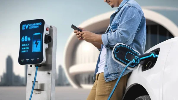 Asian man waiting and using smartphone while his electric car recharging energy from future home charging station. Smart and futuristic home energy infrastructure. Peruse