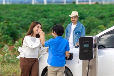 İlerici mutlu aile kavramı elektrikli araçla rüzgar çiftliğinde eğleniyor. Rüzgar türbini jeneratöründen gelen temiz yenilenebilir enerji ile şarj edilen elektrikli araç..