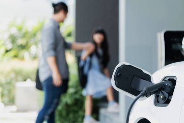 Arka planda bulanık baba ve kızının yürüdüğü şarj istasyonunda elektrikli araba şarj etmeye odaklan. Sağlıklı çevre yaşam tarzı konsepti için yenilikçi yeşil ve temiz enerji aracı.