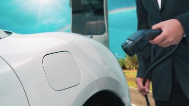 İlerici işadamı elektrikli arabayı tepe üzerindeki yolculuğunda şarj istasyonuyla dolduruyor. Temiz çevre için yenilenebilir enerji ile çalışan EV arabası. EV otomobilinden enerji sürdürülebilirliği kavramı.