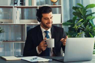Çağrı merkezi operatörünün müdürü bilgisayarla uğraşırken masasında kahvesiyle oturuyor. Kulaklık takan ve siyah takım giyen bir ofis çalışanı müşteri desteği ya da tele pazarlama üzerinde çalışıyor. coşkulu