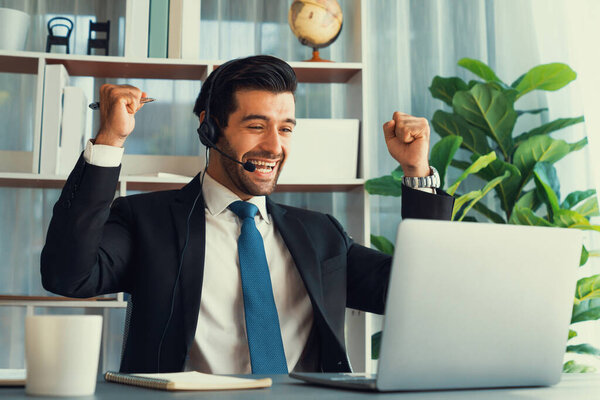 Excited and happy businessman dressed in black formal suit, raise his arm in celebratory gesture at office desk. Successfully call center or telesales operator celebrate in workplace. Fervent