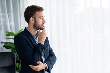 Siyah takım elbiseli yakışıklı iş adamı, modern ofis portresine güvenerek duruyor. Düşünceli bir ifadeyle iş hakkında derin düşüncelere dalmış bir sonraki hamlesini stratejik olarak düşünüyor. Varlık