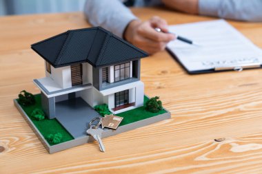 Ev modeli ahşap toplantı masasında, emlakçı ve müşterinin bulanık geçmişinde ev kredisi veya kira kontratının şartlarını tartışırken görüntülenir. Varlık