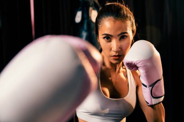 Asya 'lı kadın Muay Thai boksörü kamera önünde boksör yumruğu. Arka planda boks ekipmanıyla spor salonunda poz vermeye hazır. Kararlı gözler ve meydan okumaya hazır olun. Impetus