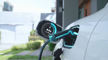 Ev şarj istasyonundaki EV şarj cihazı fişe takılı ve dijital batarya durum hologramını gösteren elektrikli arabayı şarj ediyor. Akıllı ve fütüristik ev enerjisi altyapısı. İnceleyin