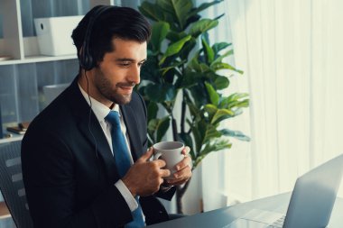 Çağrı merkezi operatörünün müdürü bilgisayarla uğraşırken masasında kahvesiyle oturuyor. Kulaklık takan ve siyah takım giyen bir ofis çalışanı müşteri desteği ya da tele pazarlama üzerinde çalışıyor. coşkulu