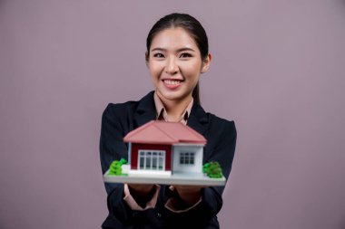 Resmi takım elbise giyen genç Asyalı kadın izole edilmiş arka planda ev modelini tutuyor ve sergiliyor. Konut ilanı, ev sigortası promosyonları ya da kişiselleştirilebilir fotokopi alanı için ideal. Coşkulu.
