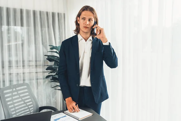 stock image Hardworking businessman stand confidently in modern office making persuasive sales call to client. Office worker talking on the phone coordinate and manage business work with colleagues. Entity