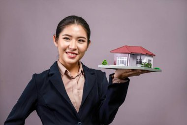 Resmi takım elbise giyen genç Asyalı kadın izole edilmiş arka planda ev modelini tutuyor ve sergiliyor. Konut ilanı, ev sigortası promosyonları ya da kişiselleştirilebilir fotokopi alanı için ideal. Coşkulu.