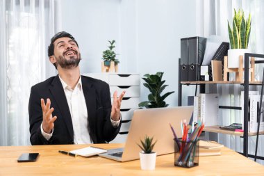 Siyah takım elbise giymiş heyecanlı ve mutlu bir iş adamı başarılı bir iş tanıtımı, enerji ve neşe kazandıktan sonra ofis masasında kutlama jesti olarak elini kaldırır. Coşkulu