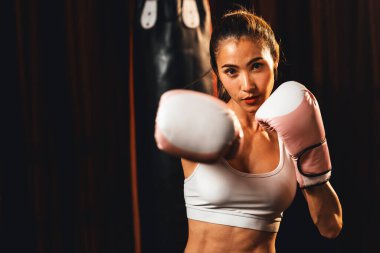 Asya 'lı kadın Muay Thai boksörü kamera önünde boksör yumruğu. Arka planda boks ekipmanıyla spor salonunda poz vermeye hazır. Kararlı gözler ve meydan okumaya hazır olun. Impetus