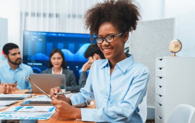 Arka plandaki ekranda bir grup ofis çalışanı ile görüşürken gözlük takan mutlu Afrikalı genç iş kadını. Takım toplantısında kendine güveni tam bir ofis hanımı. Uyum
