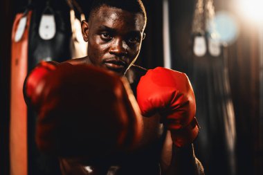 Üstsüz boksör pozu veren Afro-Amerikan Siyah Boksör yumruğunu agresif bir şekilde yumrukladı ve arka planda kum torbası ve boks ekipmanıyla spor salonunda dövüşmeye hazırdı. Impetus