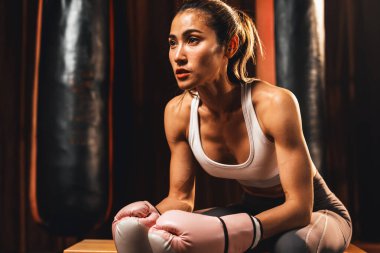 Asyalı kadın Muay Thai boksörü ya da kickboks kısa bir aradan sonra eldivenlerini giymiş ve arka planda boks ekipmanları ile salonda oturuyor. Güçlü ve kaslı vücut sporcusu. Impetus