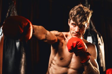 Üstsüz boksör pozu veren beyaz boksör, kameranın önünde agresif bir duruşta yumruğunu yumrukluyor ve arka planda kum torbası ve boks ekipmanıyla spor salonunda dövüşmeye hazır. Impetus
