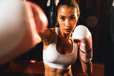Asya 'lı kadın Muay Thai boksörü kamera önünde boksör yumruğu. Arka planda boks ekipmanıyla spor salonunda poz vermeye hazır. Kararlı gözler ve meydan okumaya hazır olun. Impetus