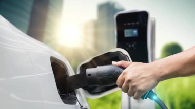Adam elektrikli araca akıllı EV şarj cihazı takıyor ve arka planda yeşil şehir parkı olan EV şarj istasyonundan şarj oluyor. ESG şehrinde gelecekteki sürdürülebilir güç ve EV araba kullanımı. İnceleyin