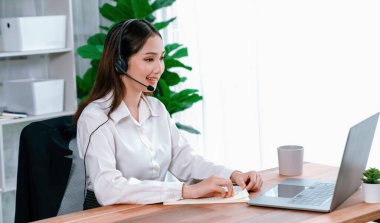 Bilgisayarında mikrofon ve kulaklıkla Asyalı çağrı merkezi çalışıyor. Kadın operatör istisnai müşteri hizmetleri sunar. Destek veren çağrı merkezi ajanı, müşteriye soruşturmada yardım ediyor. Coşkulu.