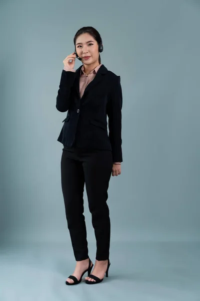 Attractive full body asian female call center operator with happy face advertises job opportunity on empty space, wearing formal suit and headset on customizable isolated background. Enthusiastic