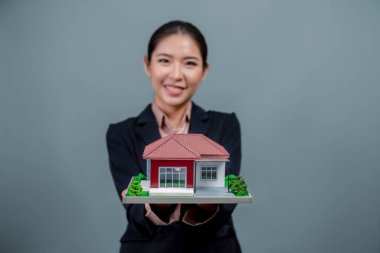 Resmi takım elbise giyen genç Asyalı kadın izole edilmiş arka planda ev modelini tutuyor ve sergiliyor. Konut ilanı, ev sigortası promosyonları ya da kişiselleştirilebilir fotokopi alanı için ideal. Coşkulu.