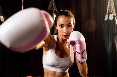 Asya 'lı kadın Muay Thai boksörü kamera önünde boksör yumruğu. Arka planda boks ekipmanıyla spor salonunda poz vermeye hazır. Kararlı gözler ve meydan okumaya hazır olun. Impetus
