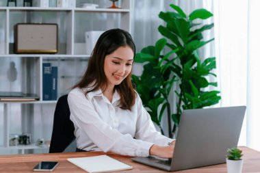Modern ofis masasındaki Asyalı hevesli genç iş kadını çalışmak ve notlar yazmak için dizüstü bilgisayar kullanıyor. Çalışkan ve çekici ofis hanımı ofisinde bilgisayar defteri üzerinde çalışıyor..