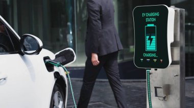 İş adamı elektrikli arabasını şehir merkezindeki şarj istasyonundan ya da otoparktan şarj ediyor. Şehir yaşam tarzında alternatif temiz enerji kullanan çevre dostu şarj edilebilir araba. İnceleyin.