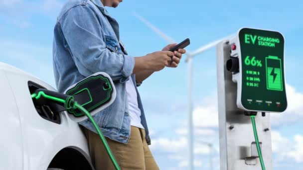 風力タービンファームの充電ステーションからEv充電器から電気自動車を充電しながらスマートフォンを使用するアジア人男性 持続可能でクリーンなエネルギー利用コンセプト パーセント — ストック動画