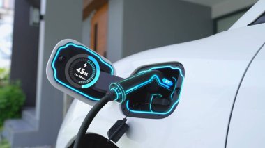 Ev şarj istasyonundaki EV şarj cihazı fişe takılı ve dijital batarya durum hologramını gösteren elektrikli arabayı şarj ediyor. Akıllı ve fütüristik ev enerjisi altyapısı. İnceleyin